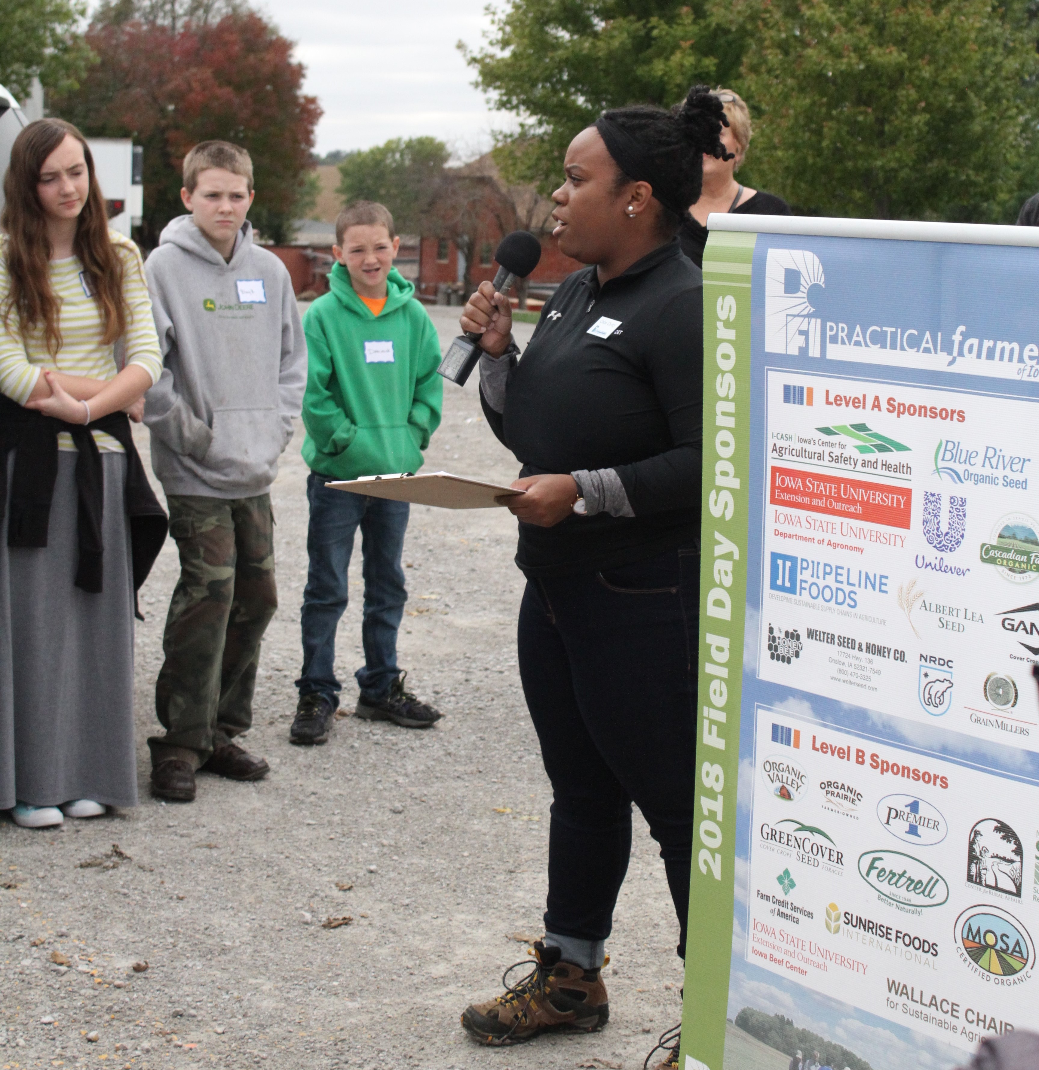 Celize Christy presents at a Practical Farmers of Iowa field day; she speaks into a microphone outside in front of an informational banner to a group of listeners.