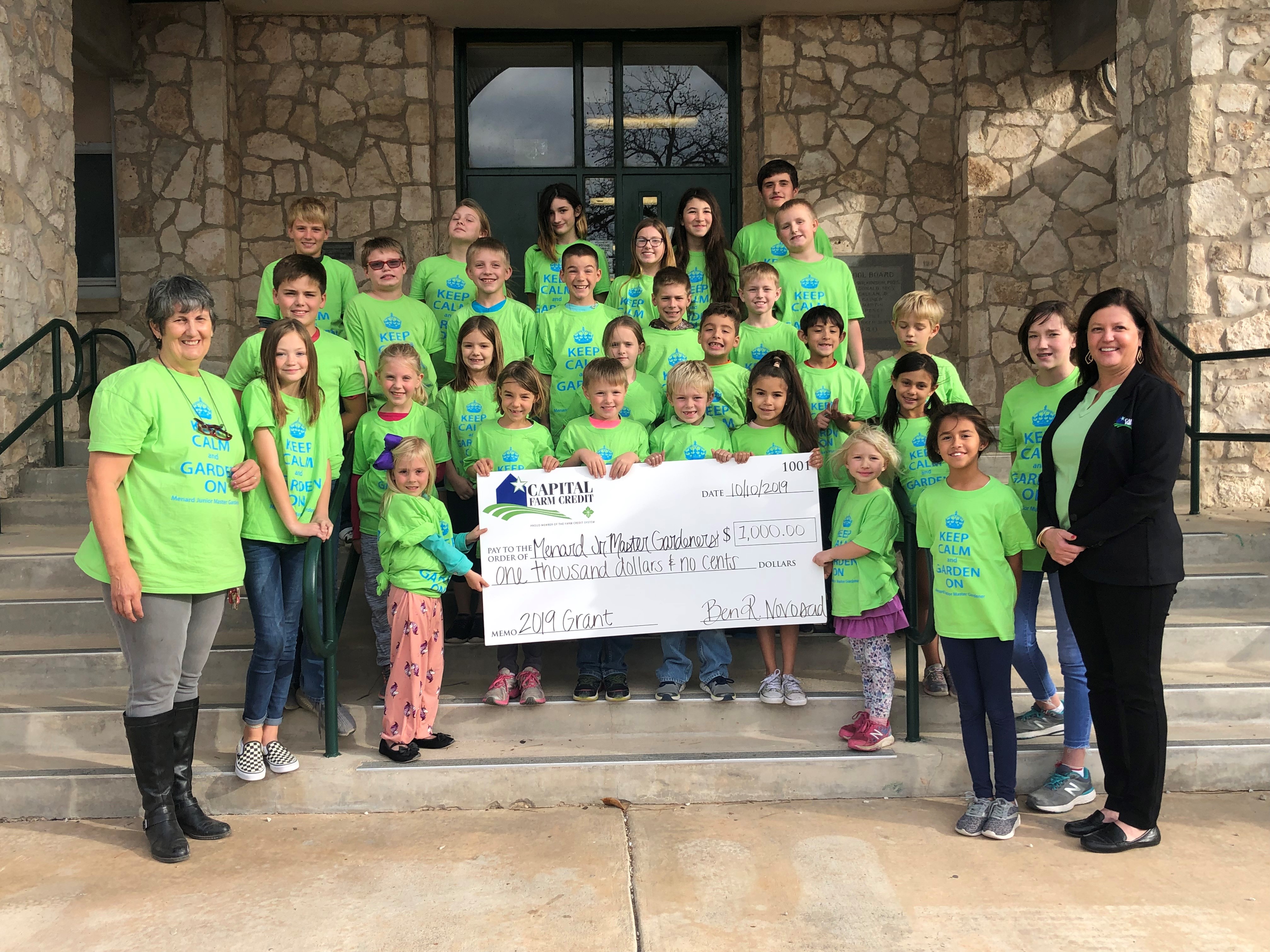 The 2019 Menard Jr Master Gardeners club stands in matching green shirts that say "keep calm and garden on" showing a check for$1,000 from Capital Farm Credit