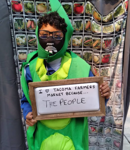 child wearing green outfit  holding sign that says they love the Tacoma Farmers Market because of the people 