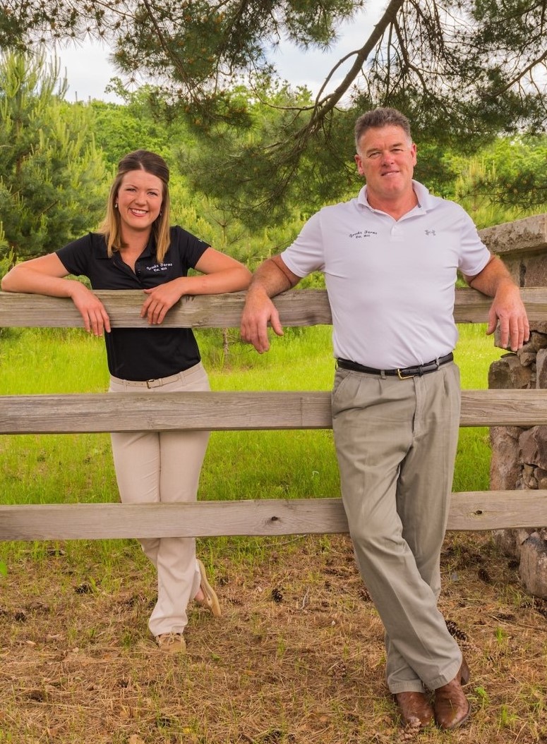 A man and woman stand against a wooden fence beneath a pine tree.