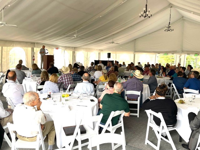 Man speaking to a crowd of maybe 50 people seated under an outdoor tent